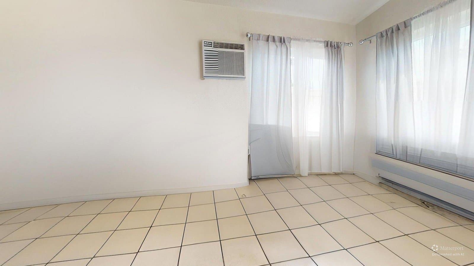 Virtual rendering: An empty living room with light-colored walls, light tile flooring, a window air conditioner, and windows covered by sheer white curtains over a baseboard heater.